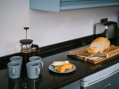 A kitchen counter with a French press coffee maker four mugs a plate of sliced melon and bread on a cutting board with a knife at Craig Cottage in Bowness-On-Windermere