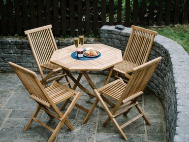 An outdoor patio with a wooden table and four wooden chairs with food and drinks on the table at Craig Cottage in Bowness-On-Windermere