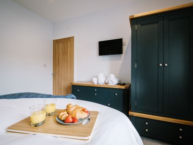 A bedroom with a tray of croissants strawberries and orange juice on a bed a black dresser with towels a black wardrobe and a wall mounted television at Craig Cottage in Bowness-On-Windermere