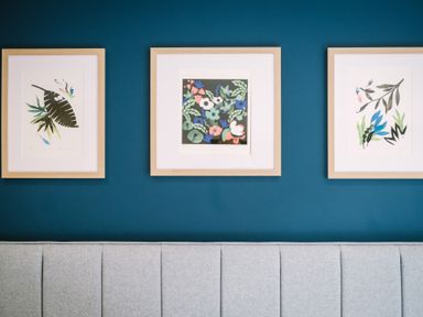 Three framed botanical prints on a blue wall above a gray upholstered headboard at Craig Cottage in Bowness-On-Windermere