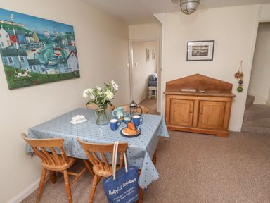 A dining area with a wooden table covered by a blue tablecloth and four wooden chairs a vase with flowers croissants on a plate and mugs on the table a wooden cabinet on the right and a painting on the left wall at 9 Harefield Cottages in Lympstone