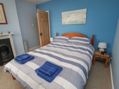 A bedroom with a double bed with blue and white striped bedding two blue towels on the bed a wooden door a nightstand with a lamp and clock on it and a small painting on a blue wall at 9 Harefield Cottages in Lympstone
