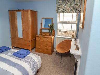 A bedroom with a bed with folded towels a wooden wardrobe and chest of drawers a wicker chair and a window with floral blinds at 9 Harefield Cottages in Lympstone