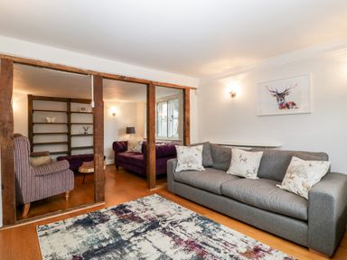 A living room with a gray sofa and a rug separated by wooden beams from another seating area with purple sofas at Oakapple Cottage in Swan Green near Lyndhurst