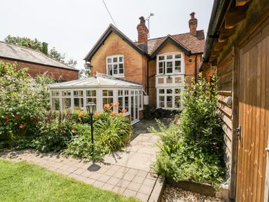 A garden with flower beds and a paved path next to a brick house and a wooden shed at Oakapple Cottage in Swan Green near Lyndhurst