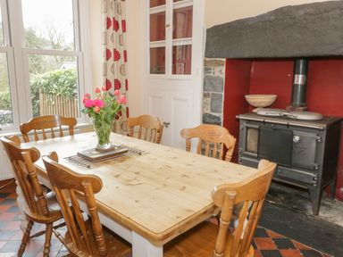 A dining room with a wooden table and chairs a vase of flowers on the table a stove in the corner and a window with curtains at Ty Capel in Betws-Y-Coed