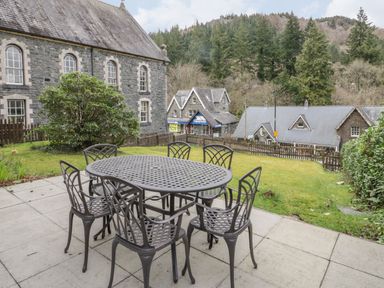 An outdoor patio with a metal table and six chairs overlooking a garden and stone buildings with trees in the background at Ty Capel in Betws-Y-Coed