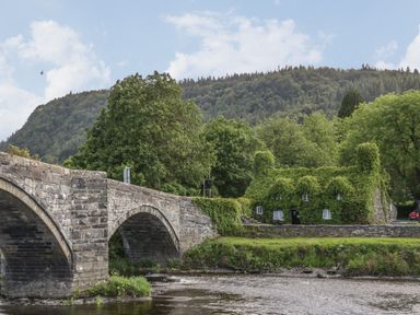 A stone bridge over a river with a house covered in ivy and trees in the background at Tyn Y Ffynnon in Betws-Y-Coed