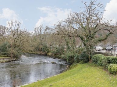 A river flowing through a wooded area with a grassy bank and a road with parked cars at Tyn Y Ffynnon in Betws-Y-Coed