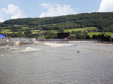 An artificial wave pool with waves and a person in the water near hills with trees and houses at Tyn Y Ffynnon in Betws-Y-Coed