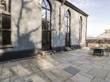 An outdoor stone patio with a table and chairs adjacent to a building with tall arched windows at Greystones Chapel in Cwm-y-Glo near Llanrug