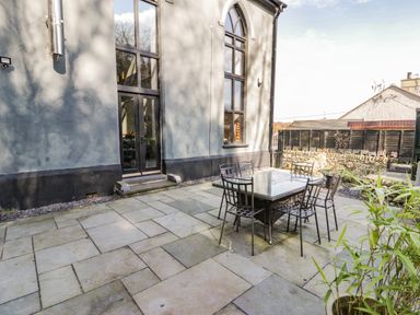 A patio area with a glass table and six metal chairs outside a building with large windows at Greystones Chapel in Cwm-y-Glo near Llanrug