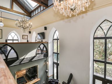 An interior view of a two-story room with arched windows chandeliers a mounted television and wooden beams at Greystones Chapel in Cwm-y-Glo near Llanrug