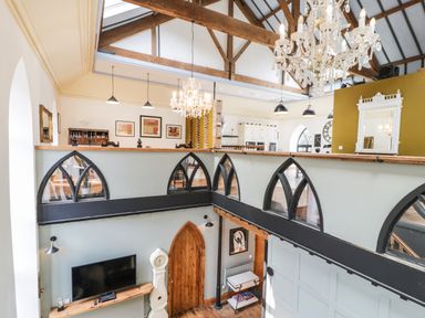 A two-story room with exposed wooden beams chandeliers a TV and a clock at Greystones Chapel in Cwm-y-Glo near Llanrug