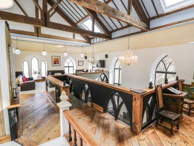 An upper floor room with wooden beams wooden floor chandeliers arched windows sitting area and wooden railing at Greystones Chapel in Cwm-y-Glo near Llanrug