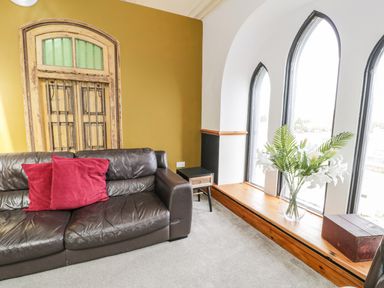 A living room with a brown leather sofa with red cushions a small side table and arched windows with a wooden ledge holding a vase of flowers at Greystones Chapel in Cwm-y-Glo near Llanrug