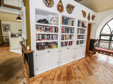 A room with white bookshelves filled with books and board games next to a wooden bench with a red cushion at Greystones Chapel in Cwm-y-Glo near Llanrug