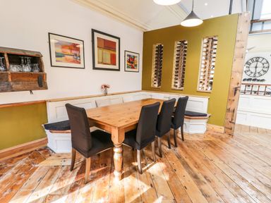 A dining area with a wooden table six black chairs built-in bench seating artwork on the wall and wooden flooring at Greystones Chapel in Cwm-y-Glo near Llanrug