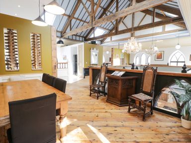 A dining area with a wooden table black chairs two wooden chairs a small cabinet chandeliers and wooden beams at Greystones Chapel in Cwm-y-Glo near Llanrug