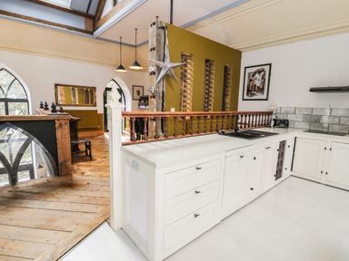 A kitchen with white cabinets and a black sink next to a wooden floor and railing with pendant lights at Greystones Chapel in Cwm-y-Glo near Llanrug