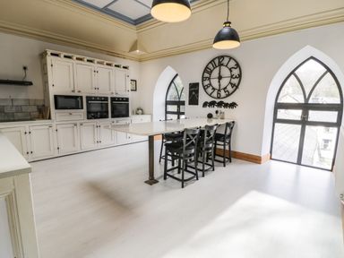 A kitchen with a large white island and black chairs next to arched windows at Greystones Chapel in Cwm-y-Glo near Llanrug