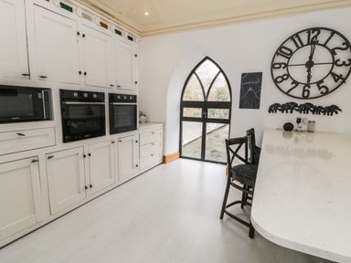 A kitchen with white cabinets black ovens a microwave a large wall clock a high countertop with black chairs and a pointed window at Greystones Chapel in Cwm-y-Glo near Llanrug