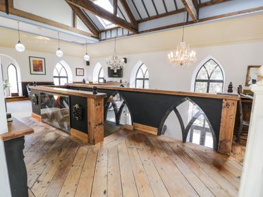 An upstairs area with wooden floors wooden railing with glass panels chandeliers and arched windows at Greystones Chapel in Cwm-y-Glo near Llanrug