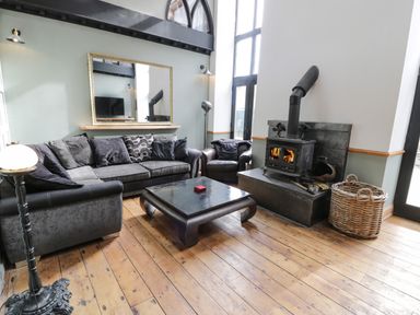 A living room with a gray sofa armchair and coffee table by a wood-burning stove with a basket of logs at Greystones Chapel in Cwm-y-Glo near Llanrug