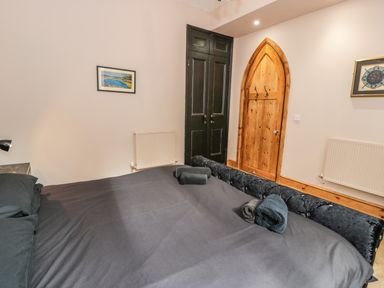 A bedroom with a black bed, rolled towels, a wooden door, a black wardrobe, radiators and framed pictures on the wall at Greystones Chapel in Cwm-y-Glo near Llanrug
