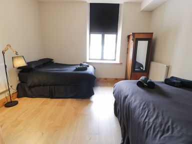 A bedroom with two beds with black bedding a wooden floor a window with a black blind a wooden wardrobe with a mirror and a floor lamp at Greystones Chapel in Cwm-y-Glo near Llanrug