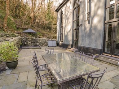 An outdoor patio with a glass-top table and metal chairs next to a building with large windows and stone walls at Greystones Chapel in Cwm-y-Glo near Llanrug