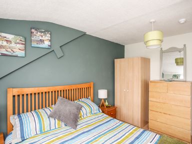 A bedroom with a wooden bed a striped bedspread a wooden wardrobe a chest of drawers and a bedside table with a lamp at Enfys in Criccieth