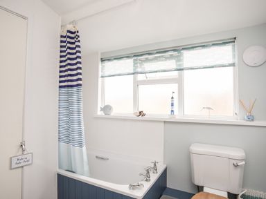 A bathroom with a bathtub that has blue paneling and a striped shower curtain a toilet with a wooden seat windows with fish-patterned blinds and small decorative items on the windowsill at Enfys in Criccieth