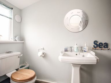 A bathroom with a toilet with a wooden seat and a pedestal sink with soap dispensers and a round mirror above at Enfys in Criccieth