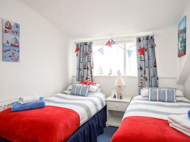 A bedroom with two single beds with striped bedding beside a window with nautical curtains and a bedside table with a lamp at Enfys in Criccieth