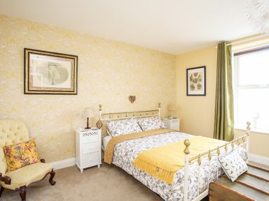 A bedroom with a bed and two bedside tables with lamps a chair with a cushion and a window with green curtains at Enfys in Criccieth