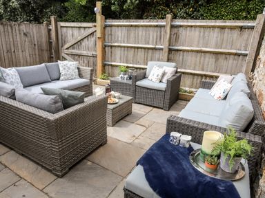 An outdoor patio with grey wicker seating and cushions a small table with tea set and snacks and a tray with plants and candles at Enfys in Criccieth