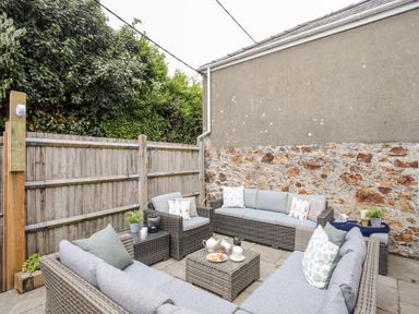An outdoor patio area with gray wicker furniture and cushions a wooden fence and stone wall at Enfys in Criccieth