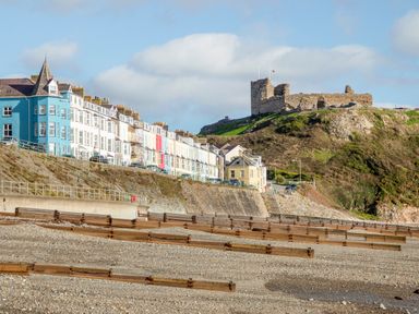 A pebble beach with wooden groynes and a row of colorful houses beside a hill with castle ruins at Enfys in Criccieth