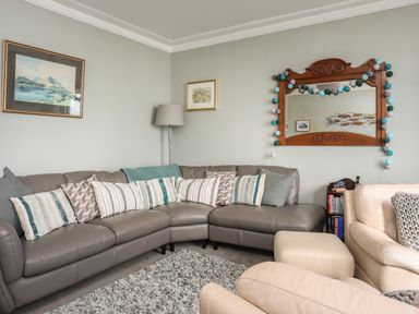 A living room with a grey corner sofa and striped cushions a beige armchair a floor lamp a wooden framed mirror and framed pictures at Enfys in Criccieth