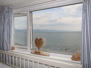 A window with blue and white striped curtains looking out over the sea with a wooden heart and fish decoration on the windowsill at Enfys in Criccieth