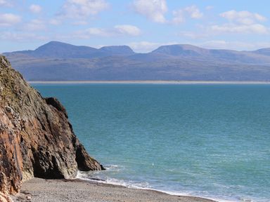 A rocky beach with a cliff on the left and a body of water with mountains in the background at Enfys in Criccieth