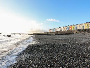 A rocky beach with waves and colored houses along the shore at Enfys in Criccieth