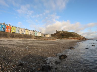 A pebble beach with colorful houses on a seawall and a hill with a castle ruin at Enfys in Criccieth