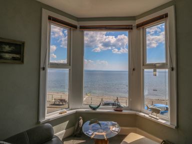 A sitting area with a glass table and decorative sea-themed items on the window sill overlooking the beach and sea at Enfys in Criccieth