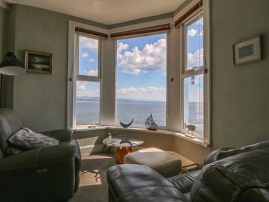 A sitting area with leather chairs and a table by bay windows overlooking water at Enfys in Criccieth