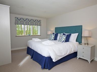 A bedroom with a double bed with a blue headboard and patterned cushions beside a window with patterned blinds and a white bedside table with a lamp at 3 The Drive Hillfield Village in Dartmouth