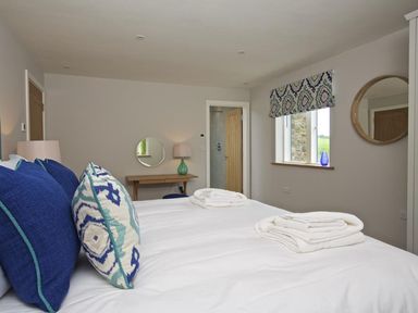 A bedroom with a bed with blue and patterned pillows two folded white towels on top a wooden door a small wooden table with a green lamp a round mirror on the wall and a window with a patterned valance at 3 The Drive Hillfield Village Dartmouth