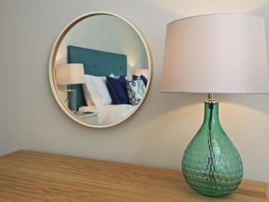 A green glass table lamp on a wooden surface with a round wall mirror reflecting a bed with blue cushions at 3 The Drive Hillfield Village in Dartmouth