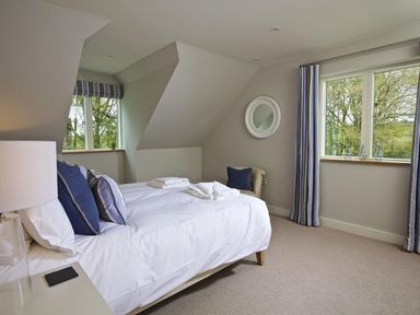 A bedroom with a bed with white bedding and blue pillows next to windows with curtains and a small chair at 3 The Drive Hillfield Village Dartmouth